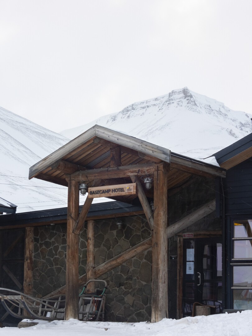Snow-covered timber entrance to Basecamp Hotel in Longyearbyen, with its hotel sign and mountains behind.