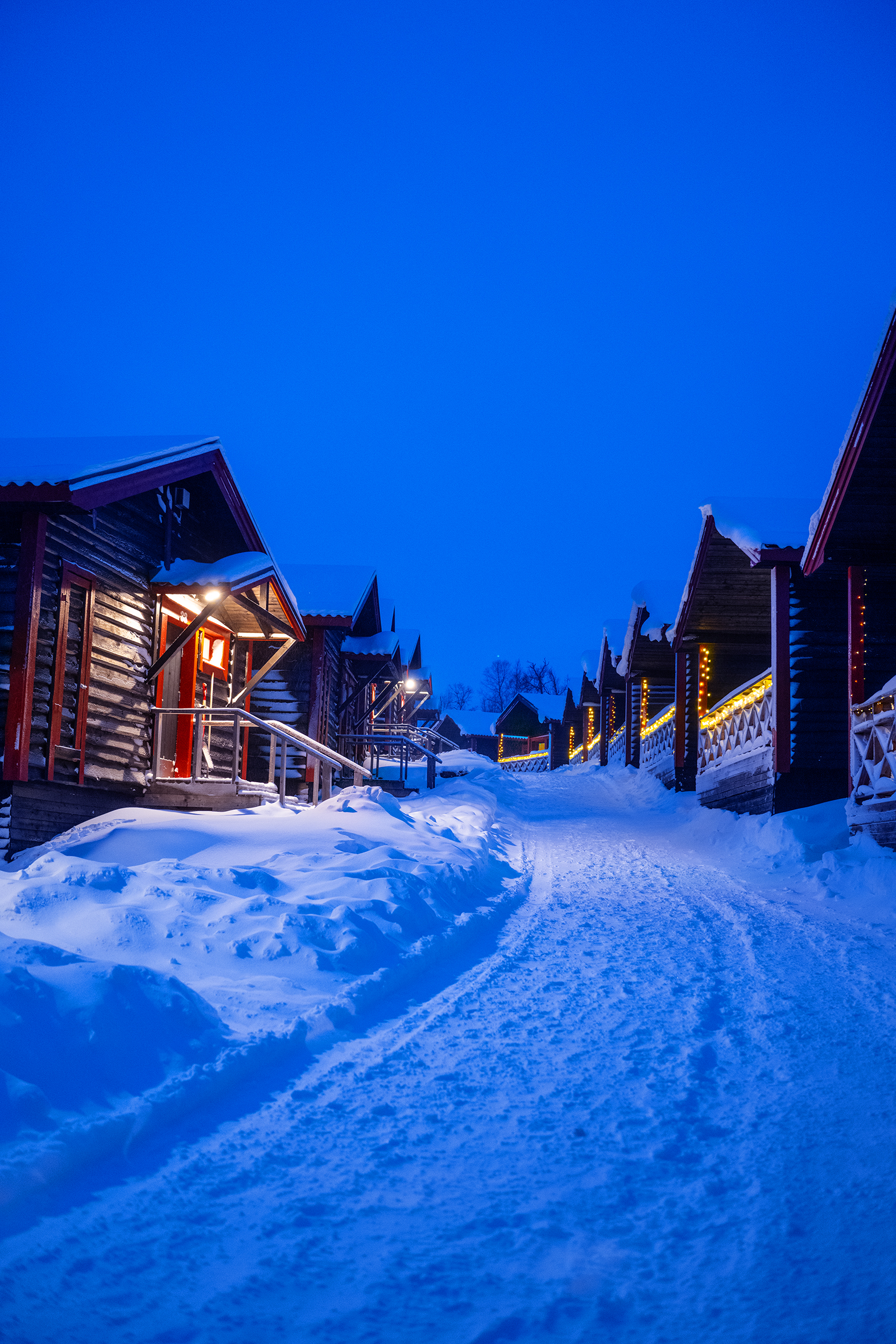 Snow-covered timber cabins with lit windows in Björkliden’s cottage village at blue hour.
