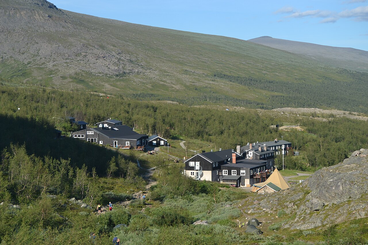 The dark timber buildings of STF Kebnekaise Mountain Station below a green mountain slope in summer.